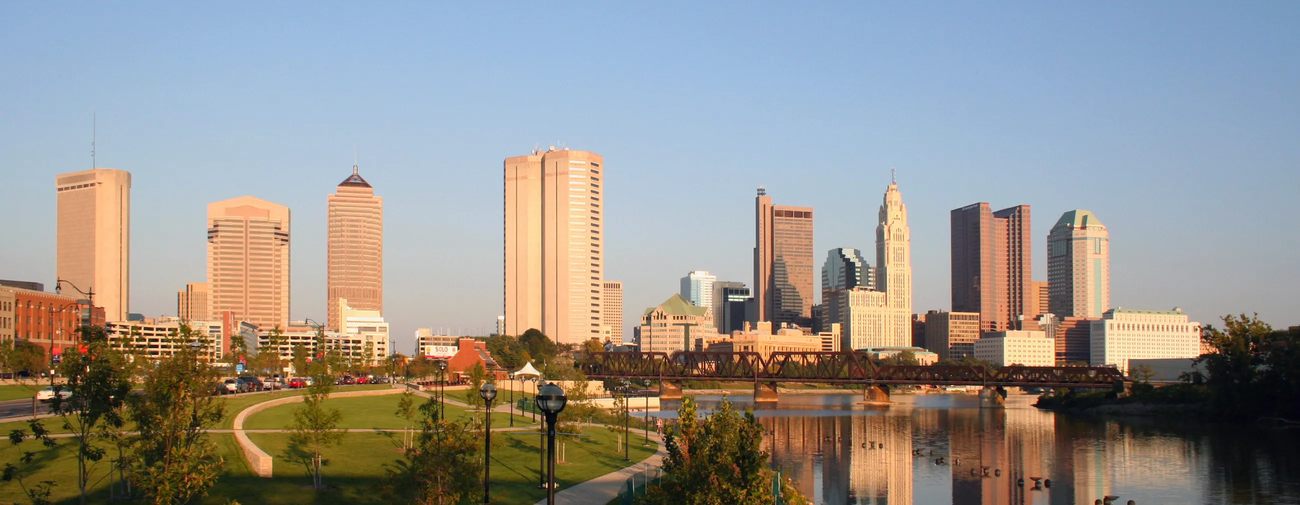 The downtown Columbus skyline in late afternoon light, seen across the Scioto River from the west bank.