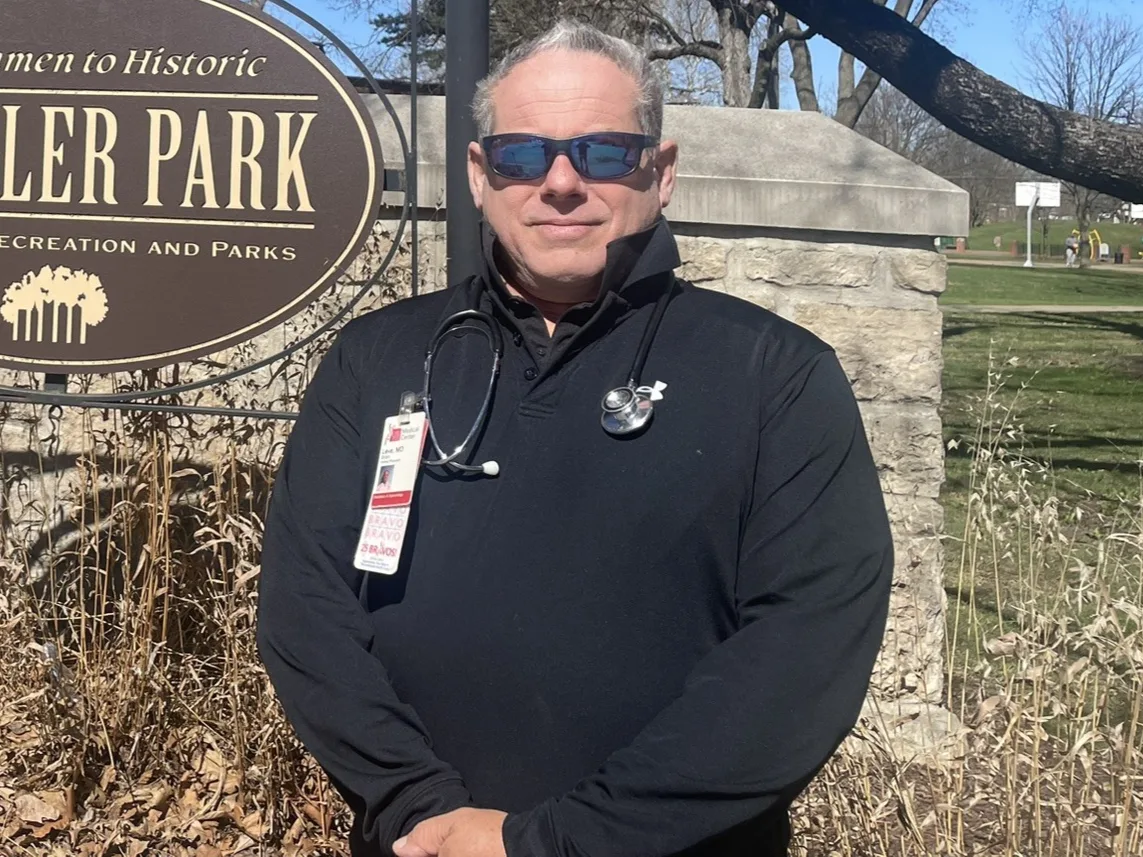 Dr. Brian Leve standing at the Welcome to Historic Schiller Park sign in German Village, in a black quarter-zip with a stethoscope and a hospital badge reading Leve, MD.