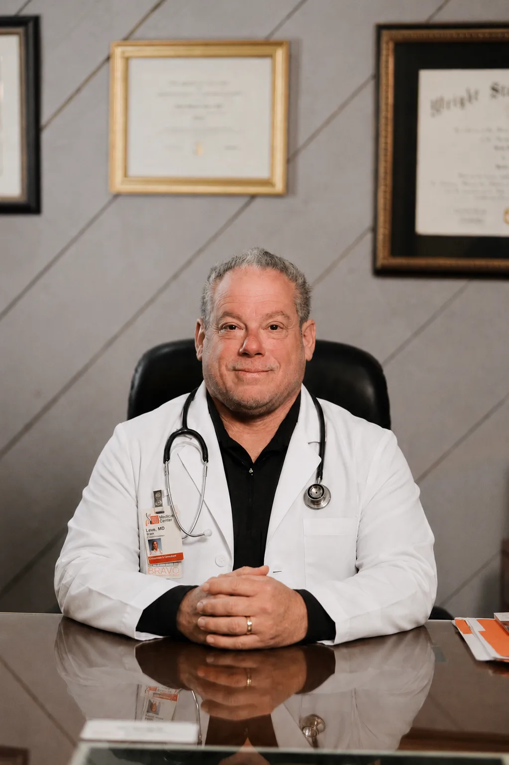 Dr. Brian Leve seated at his desk in a white coat with a stethoscope and hospital badge, framed medical diplomas on the wall behind him.